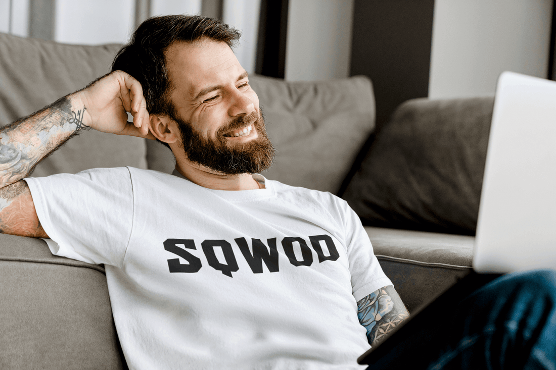 Smiling man in a SQWOD t-shirt sitting on a couch and looking at a laptop